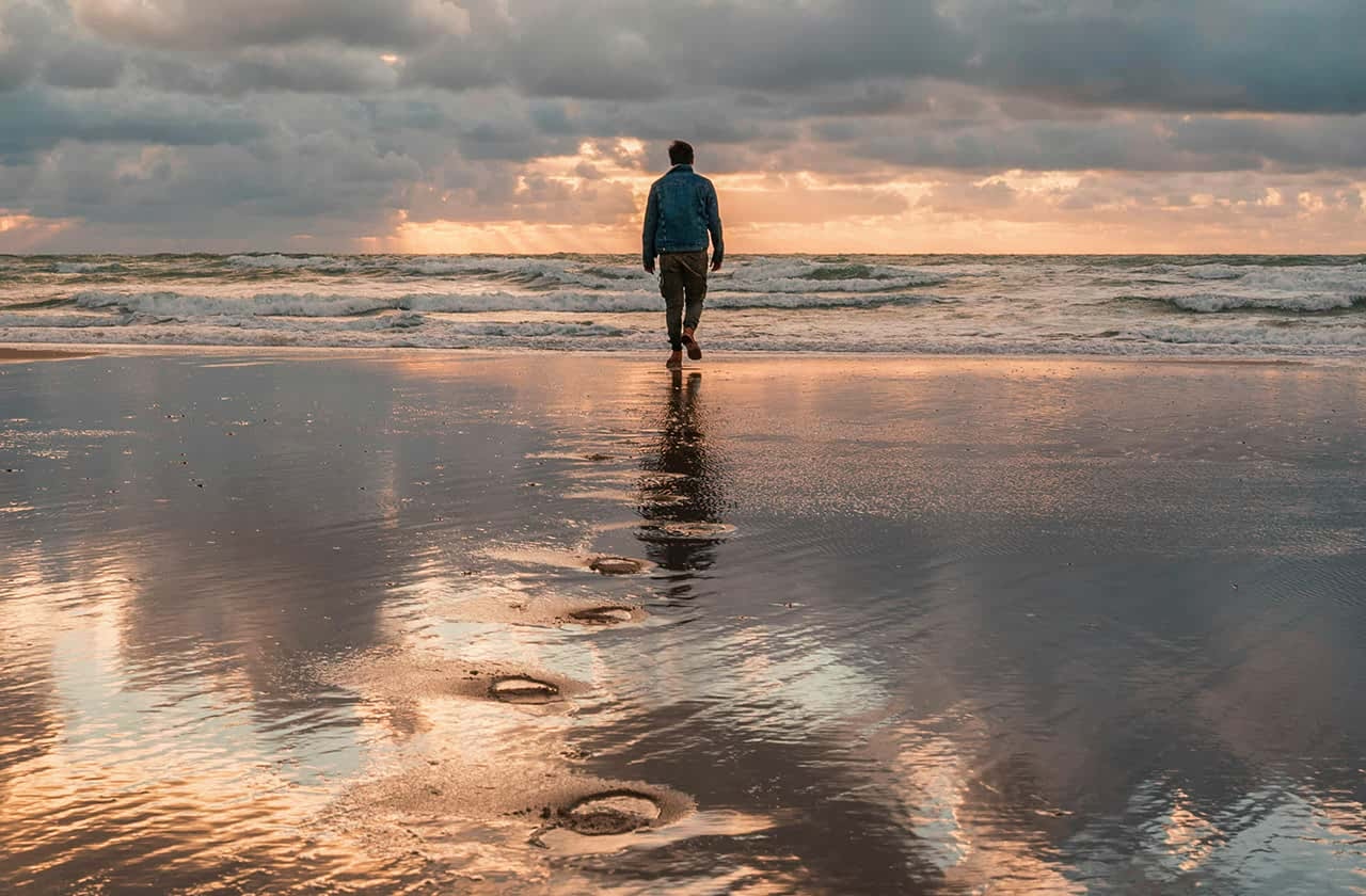 Eine Person läuft im Abendlicht auf einem Sandstrand Spuren hinterlassend auf das Wasser zu